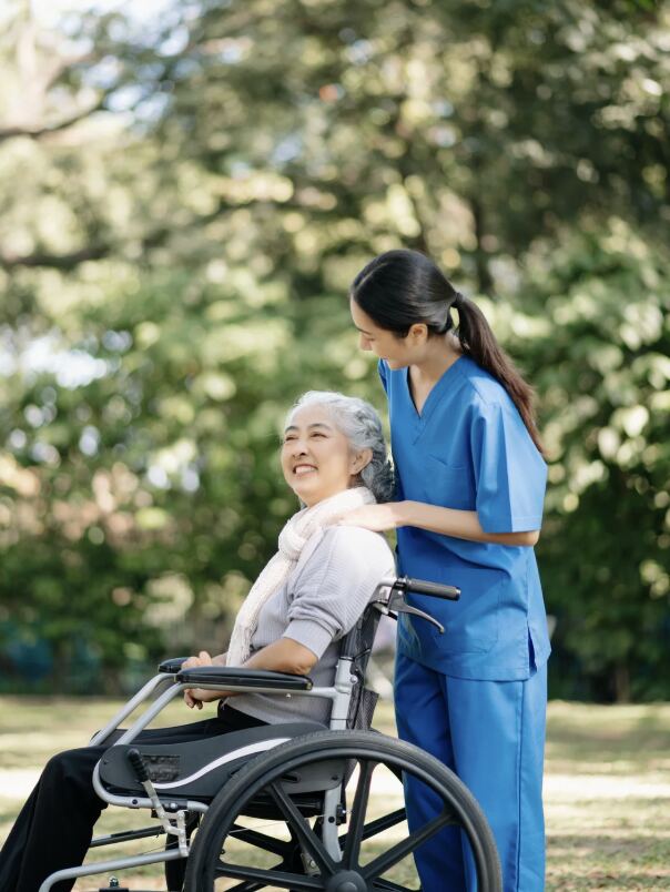 Pflegekraft begleitet eine lächelnde Seniorin im Rollstuhl im Park